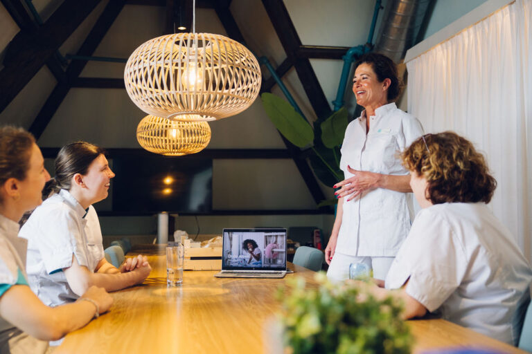 Groep collega's zit aan lunchtafel terwijl één collega vanaf haar laptop iets presenteert aan de groep
